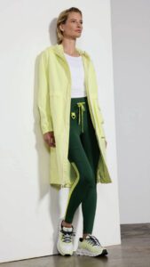 Woman standing in a studio, wearing a yellow jacket, white shirt, green pants, and sneakers, looking to the side.