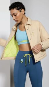 A woman in fashionable activewear, consisting of blue leggings and a crop top, stylishly wears a light beige jacket and a lime green inner lining.