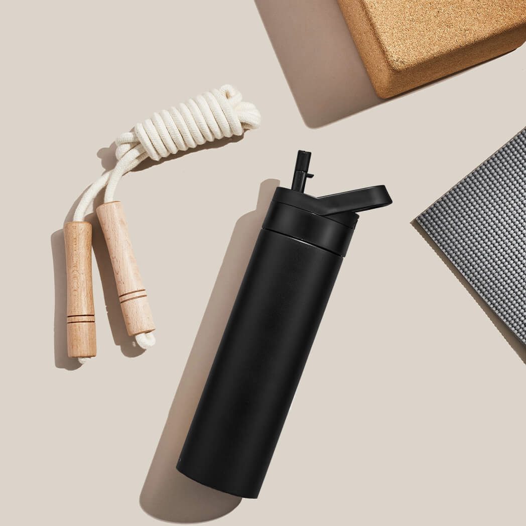 A black water bottle with a drinking straw beside a skipping rope and a cork yoga block on a beige surface.