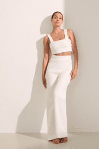 Woman in a white crop top and pants standing against a light-colored wall, casting a soft shadow.