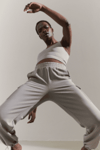 A woman in a white tank top and gray sweatpants striking a dynamic dance pose on a plain background.