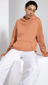 Woman in a peach hoodie and white pants sitting on a white block against a gray background.