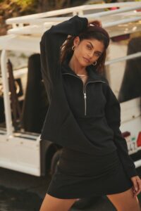 A woman in a black hoodie and skirt poses confidently beside a van, touching her hair and looking at the camera.