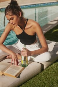 A young woman reads a book by a poolside, holding a bottle of beverage, dressed in a black tank top and white pants.