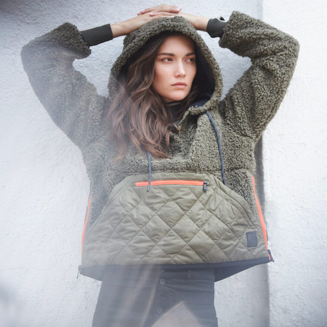 A woman in a textured green jacket with a quilted panel leans against a white wall, looking to the side thoughtfully.