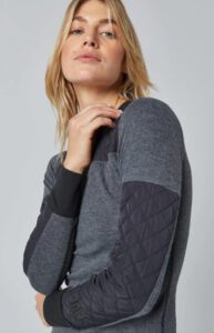 A woman with blonde hair poses in a layered gray top with quilted elbow patches, looking at the camera.